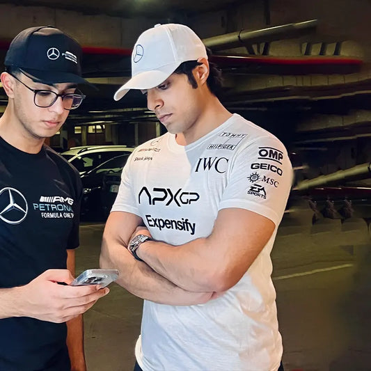 Two men in a garage, one wearing a black shirt with Mercedes-Benz logo and the other in a white shirt with various sponsor logos.