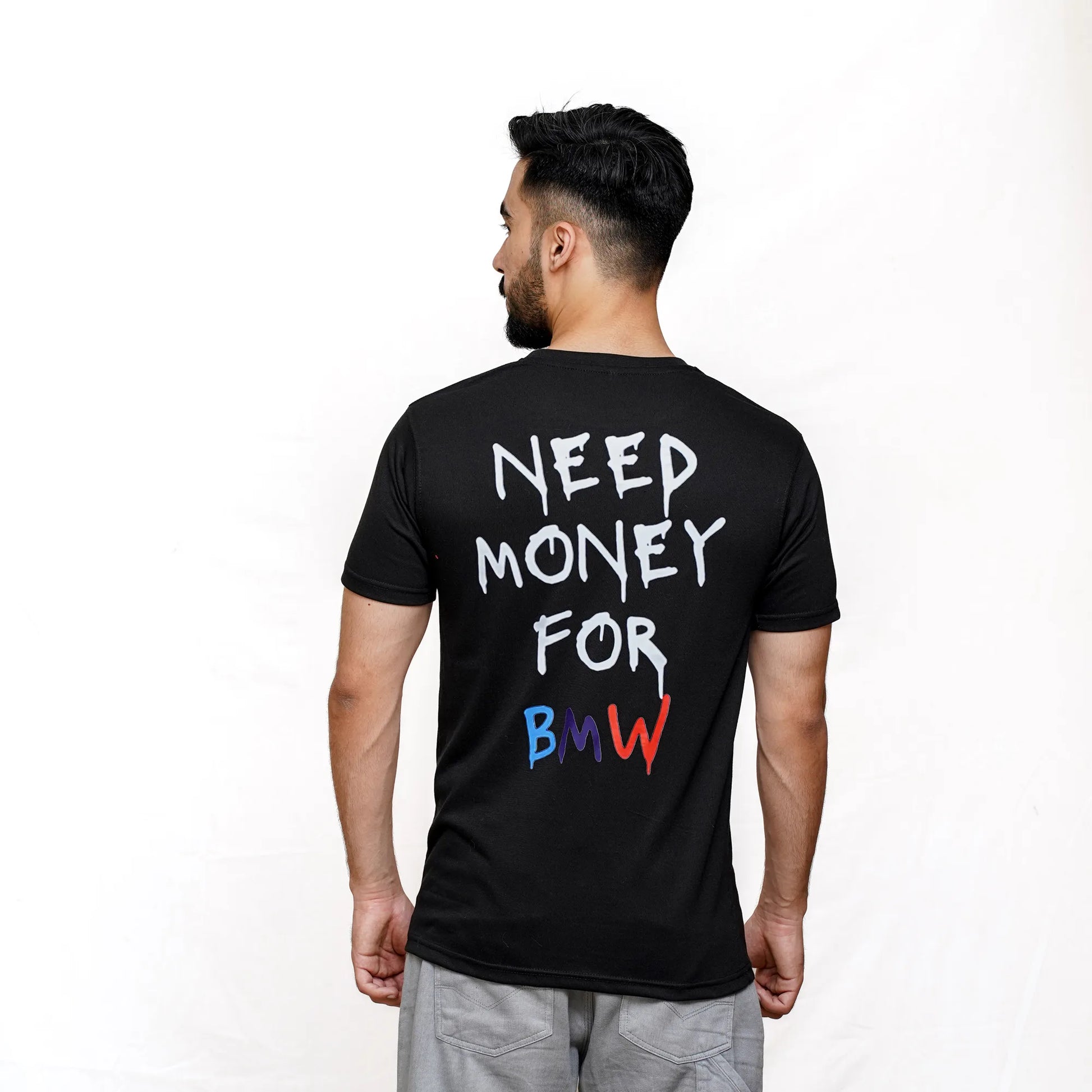 A person facing away from the camera wears a black BMW-themed t-shirt with the text 'NEED MONEY FOR BMW' printed on the back in bold, multicolored letters.