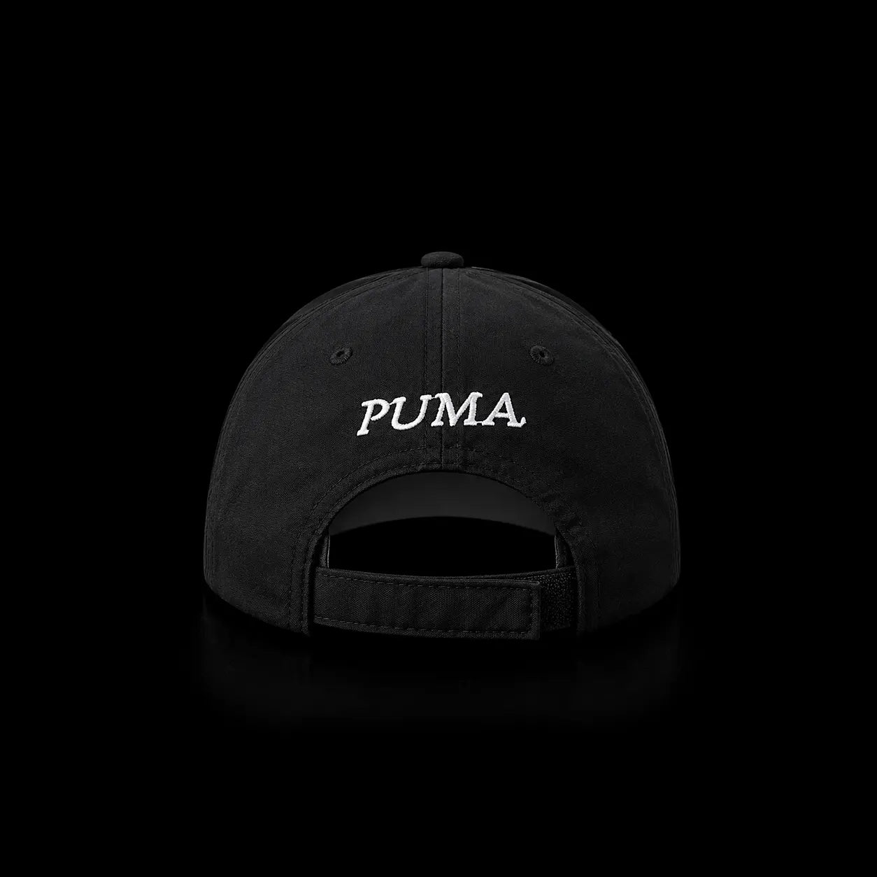 A black baseball cap shown from the back, featuring the word 'PUMA' embroidered in white script across the center. The cap is photographed against a solid black background with a subtle reflection below.